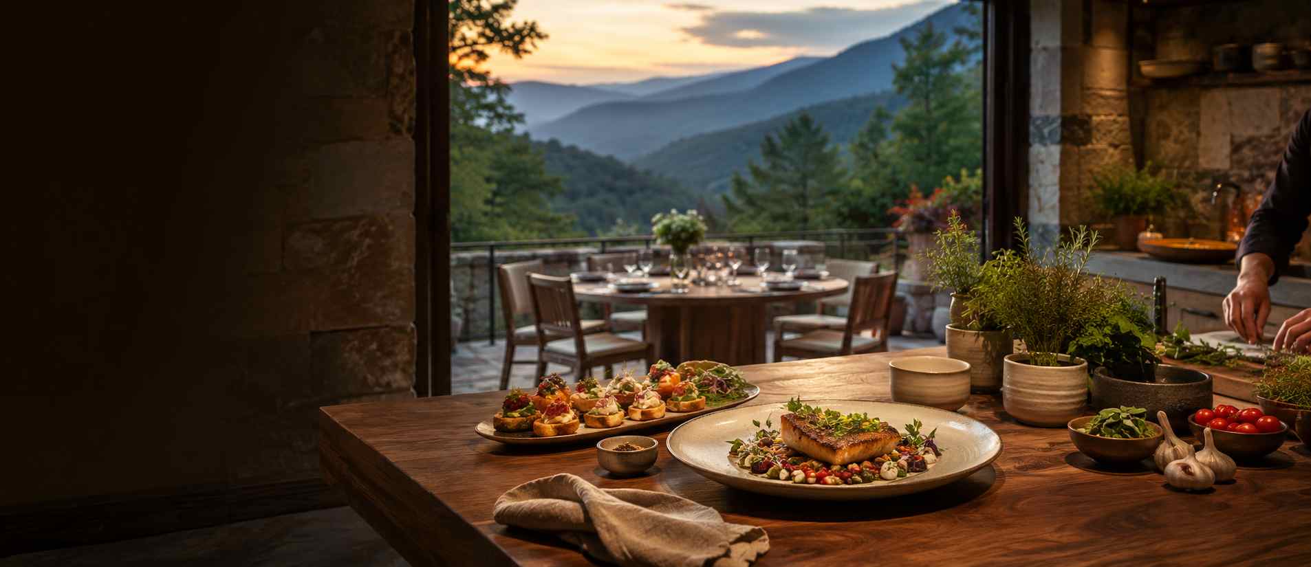 Wooden kitchen island with plated appetizers and a main dish, herb pots, and a mountain-view terrace at sunset.