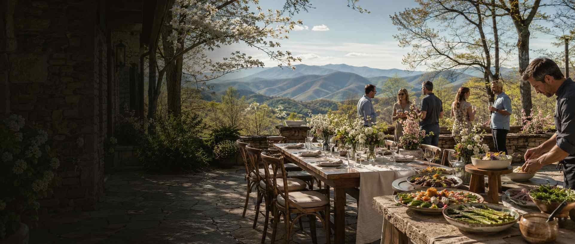Outdoor mountain-view terrace with a long dining table set for a feast; guests chat as a chef arranges dishes in the foreground.