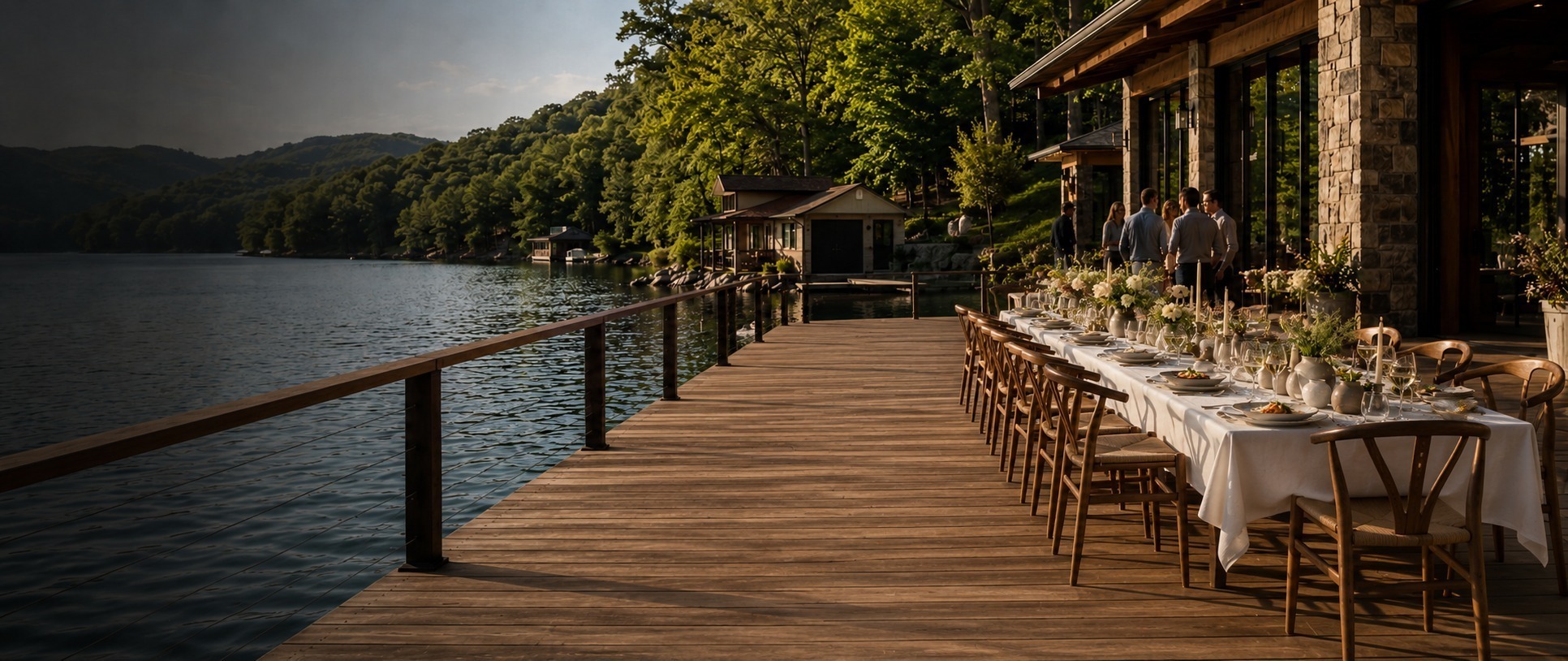 Lakeside wooden dock with a long banquet table set for an outdoor dinner, candles and floral centerpieces.