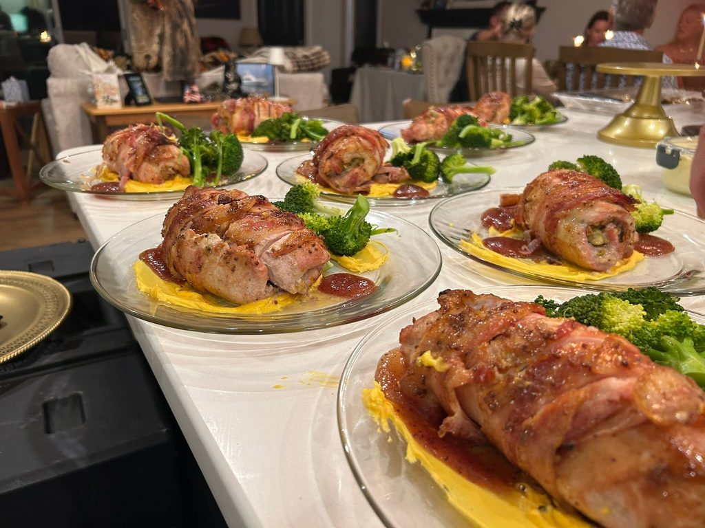 Bacon-wrapped meat portions on yellow mashed potatoes with broccoli on a banquet table of plated dishes.