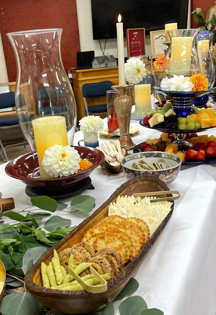 Banquet table with candles, white flowers, and a long wooden tray of crackers and pastries surrounded by greenery and fruit