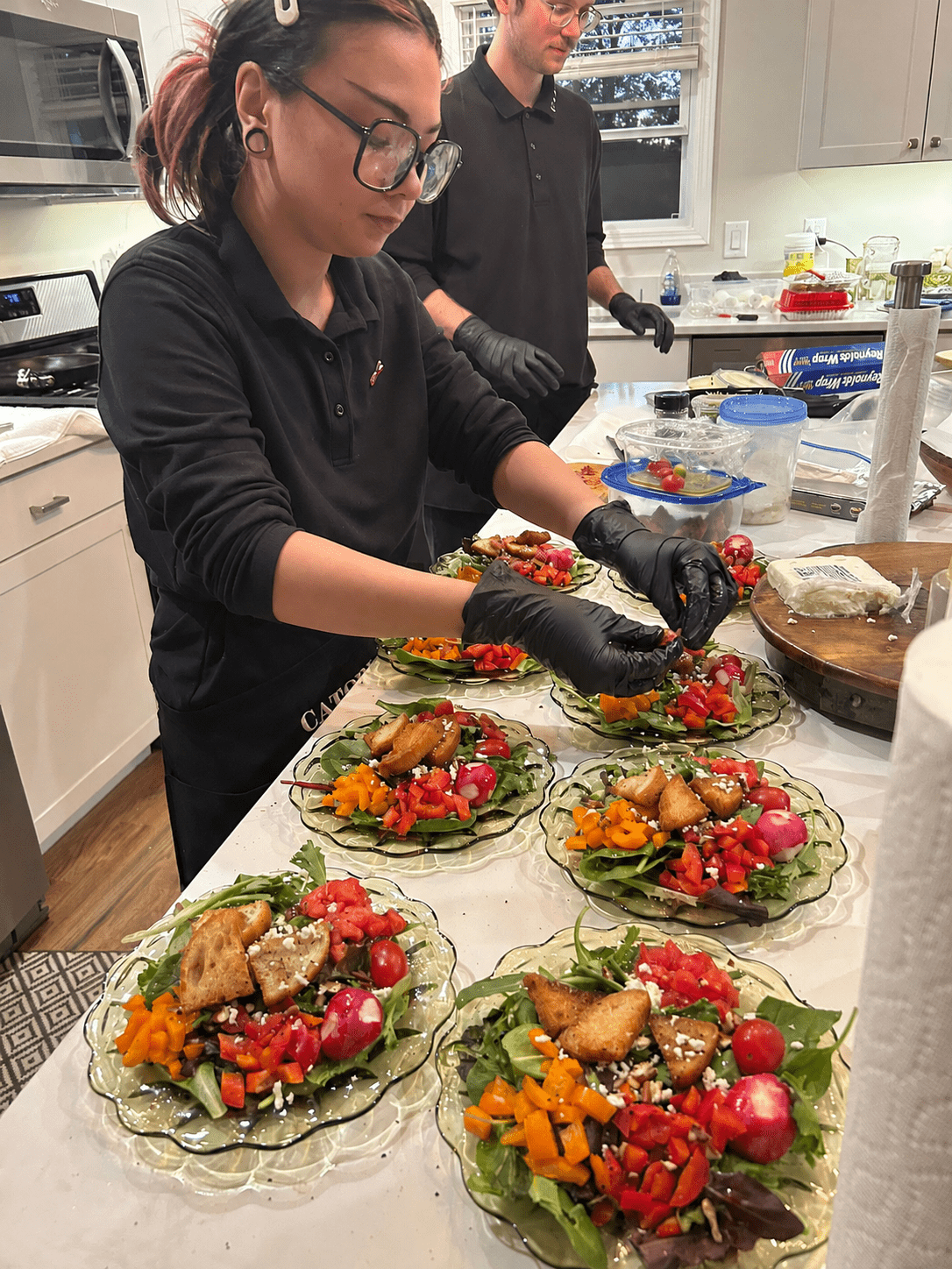 Two people in a bright kitchen garnish colorful salads on glass plates, wearing black outfits and gloves while arranging ingredients at a prepping station.