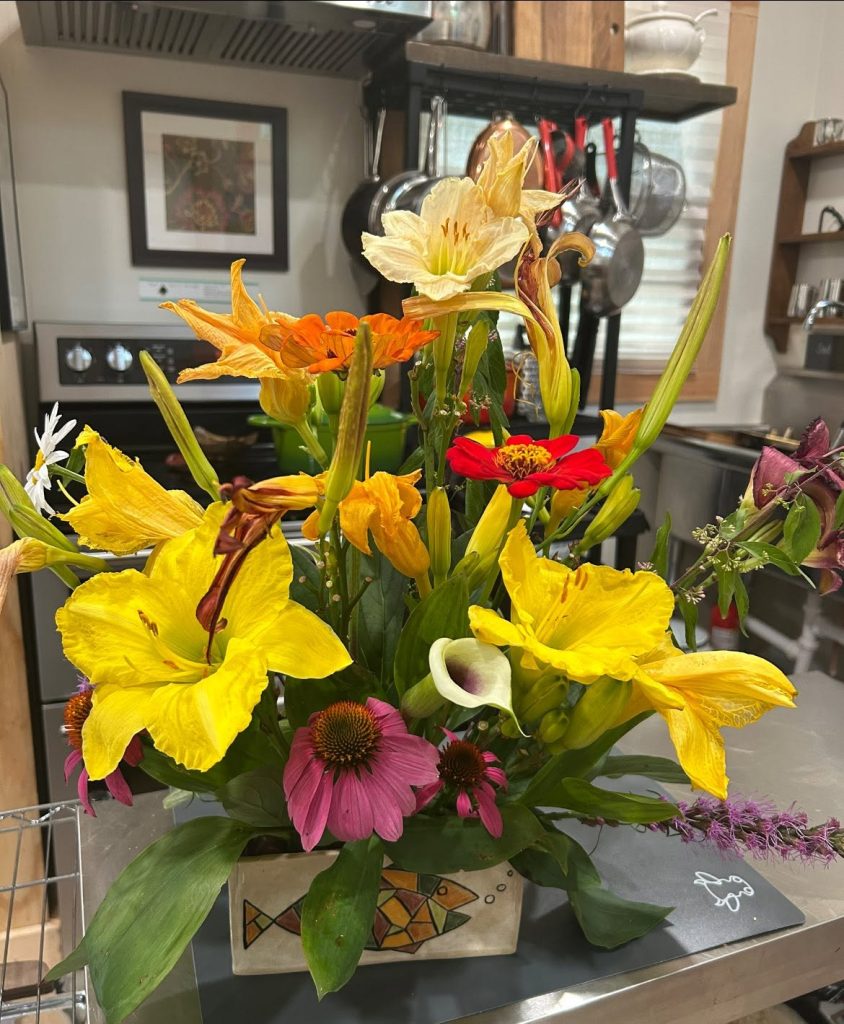 Colorful bouquet of yellow irises, orange lilies, pink coneflowers, and red zinnia in a ceramic vase on a kitchen counter.