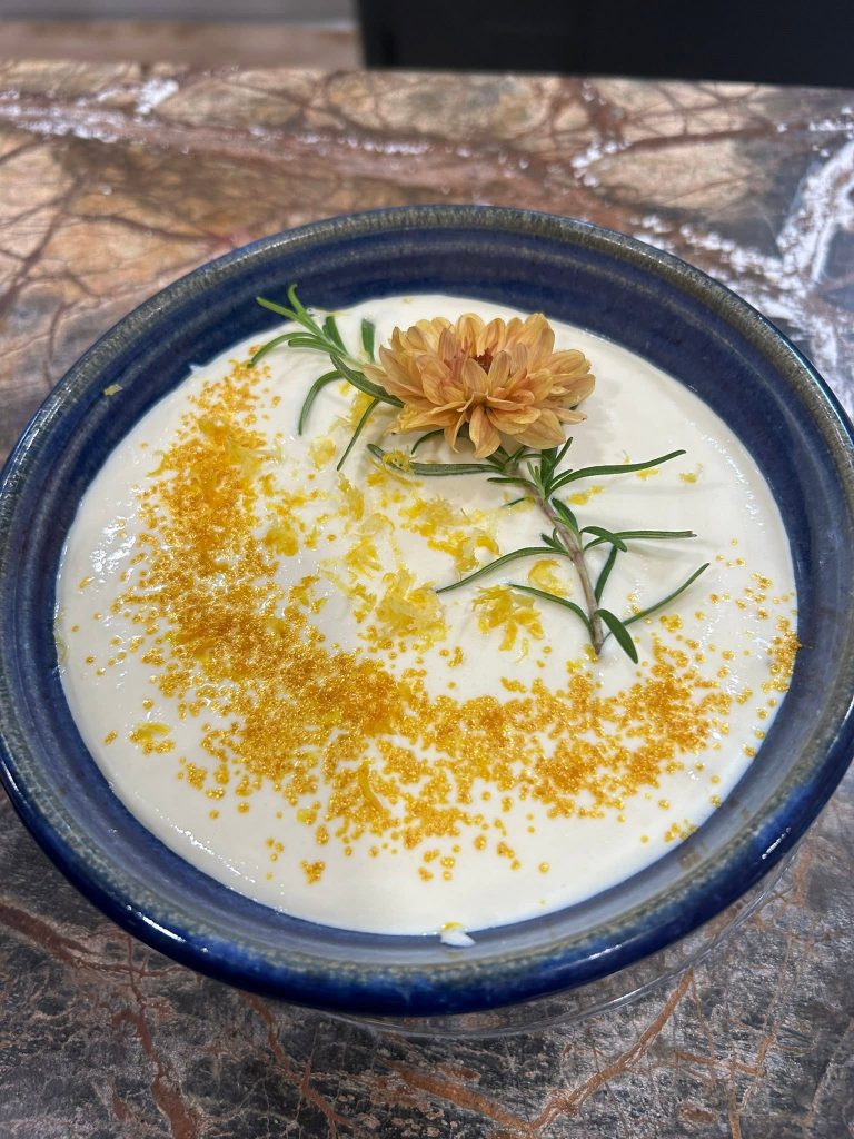 Catori Artisan Creamy Soup With Edible Flower - CATORI Artisan Blue ceramic bowl with creamy white dessert, topped with orange zest, a rosemary sprig, and a dried orange flower on a marble surface.