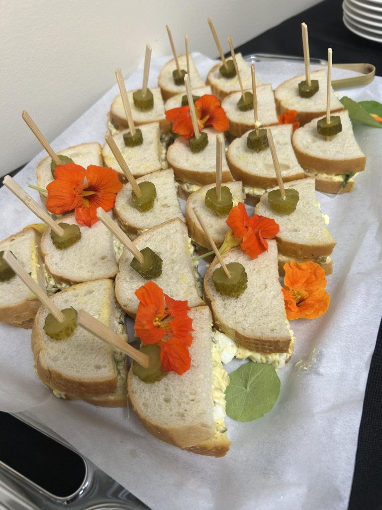 Platter of bite-size egg-salad sandwiches topped with olives and secured with toothpicks, accented by orange edible flowers and green leaves.