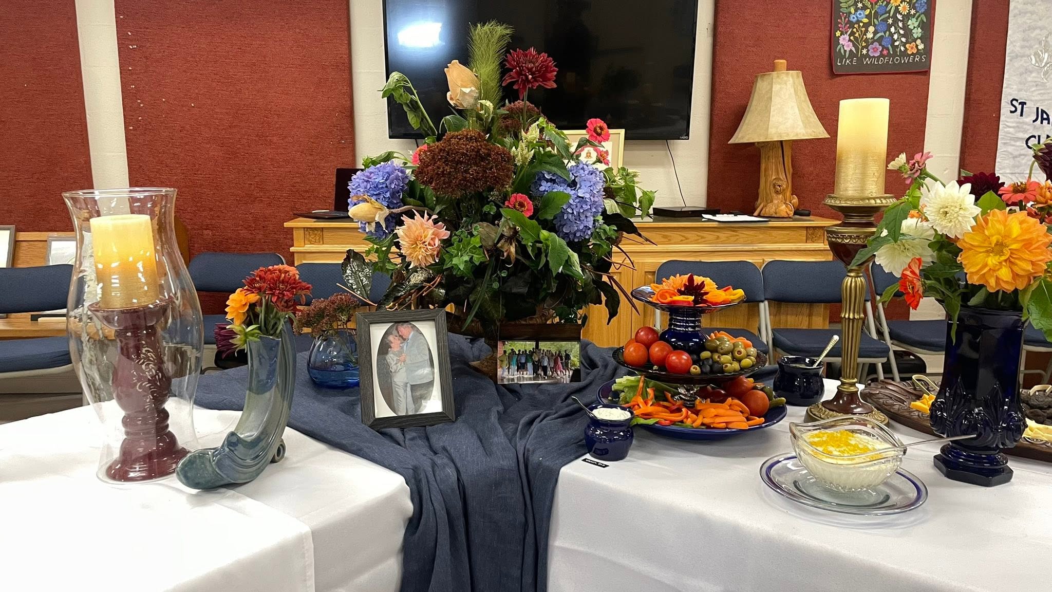 Table centerpiece for a memorial display featuring a large flower arrangement, framed photos, candles, and a fruit platter against a red wall backdrop in a room setup.