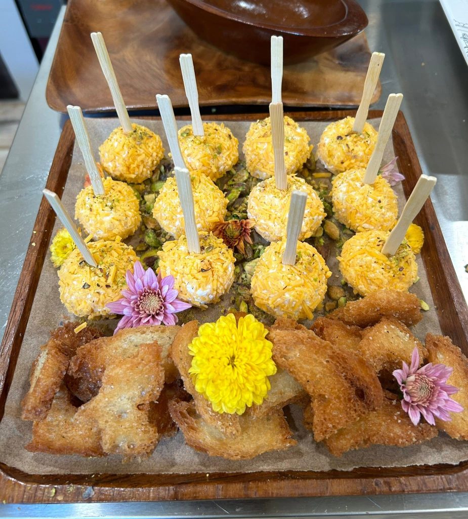 Assorted yellow-crusted fried bite-sized balls on a wooden tray with light wooden skewers, garnished with yellow and purple flowers.
