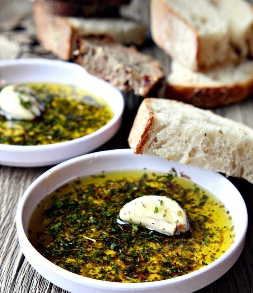 White bowls of herb-infused olive oil dip with garlic and chopped green herbs, accompanied by slices of crusty bread on a wooden table, ready for dipping.