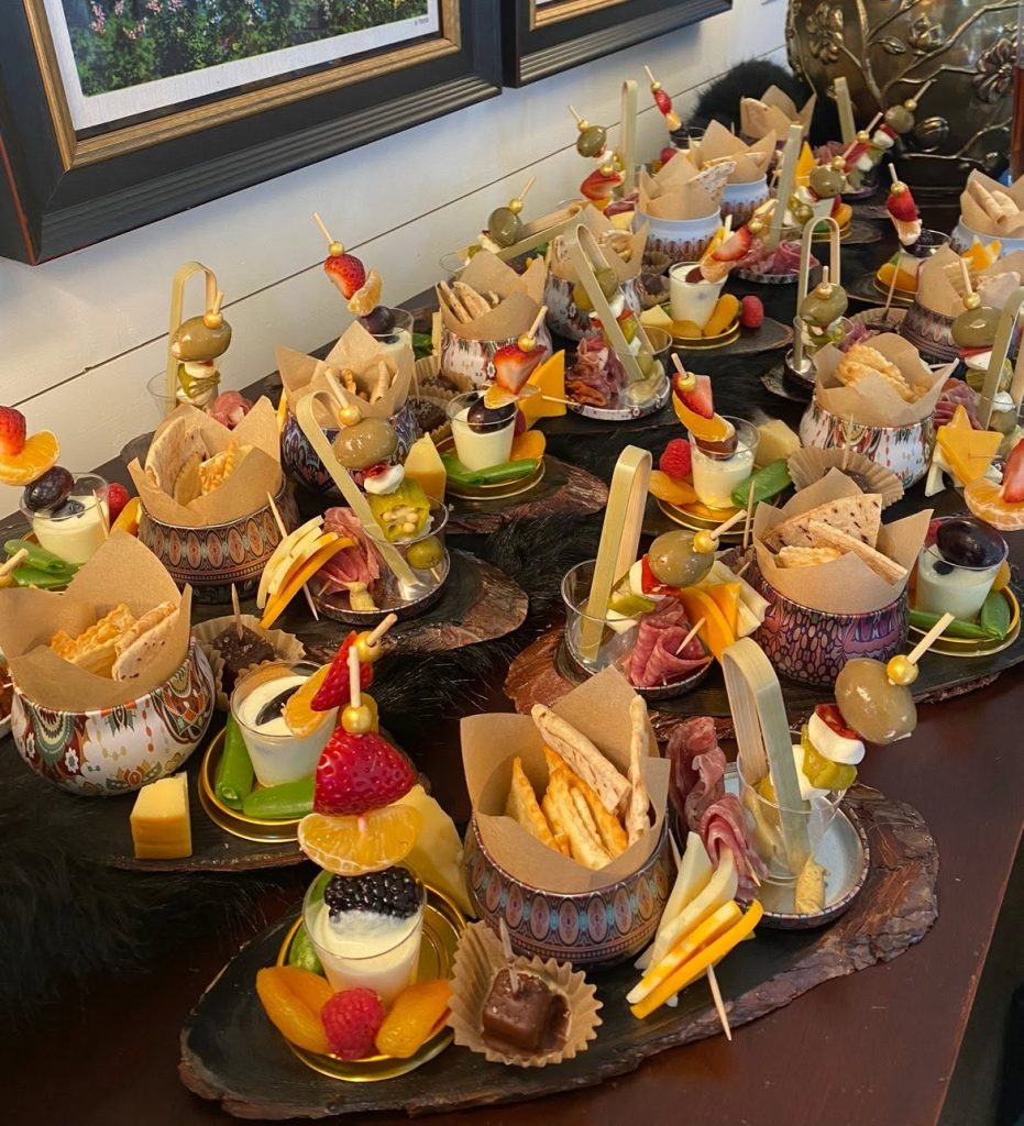 Assorted bite-sized hors d'oeuvres arranged in decorative bowls and skewers on a wooden table, with olives, cheese, fruit, and crackers.