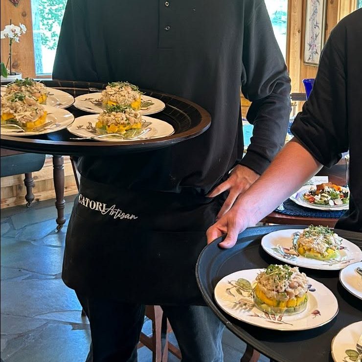 Catori Artisan Servers Presenting Plated Salads - CATORI Artisan Two male servers in black polo shirts carry circular trays with multiple plated appetizers in a sunlit wooden dining room.