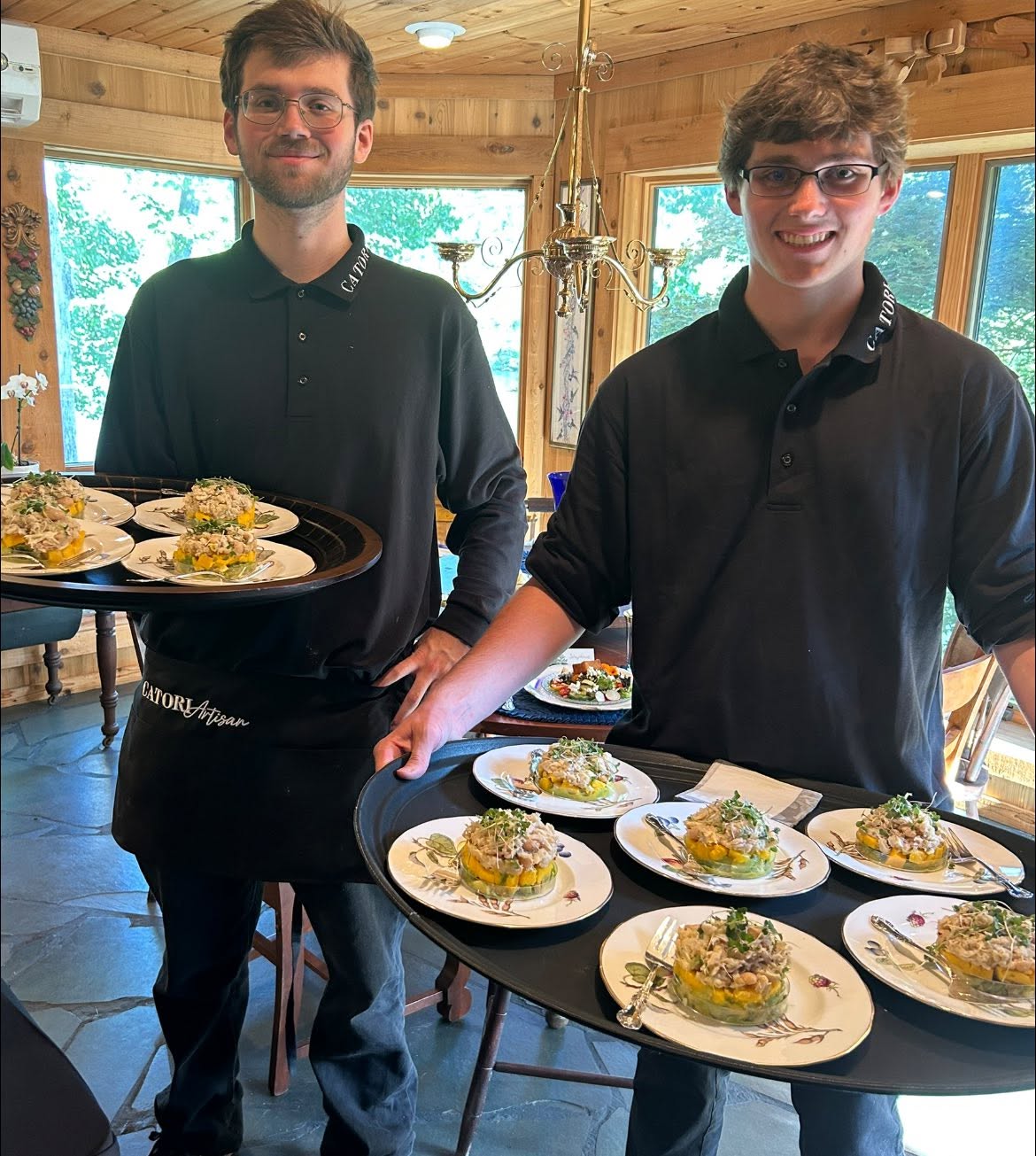 Two servers in black polo shirts carry round trays of garnished bite-size appetizers in a wood-paneled dining room.