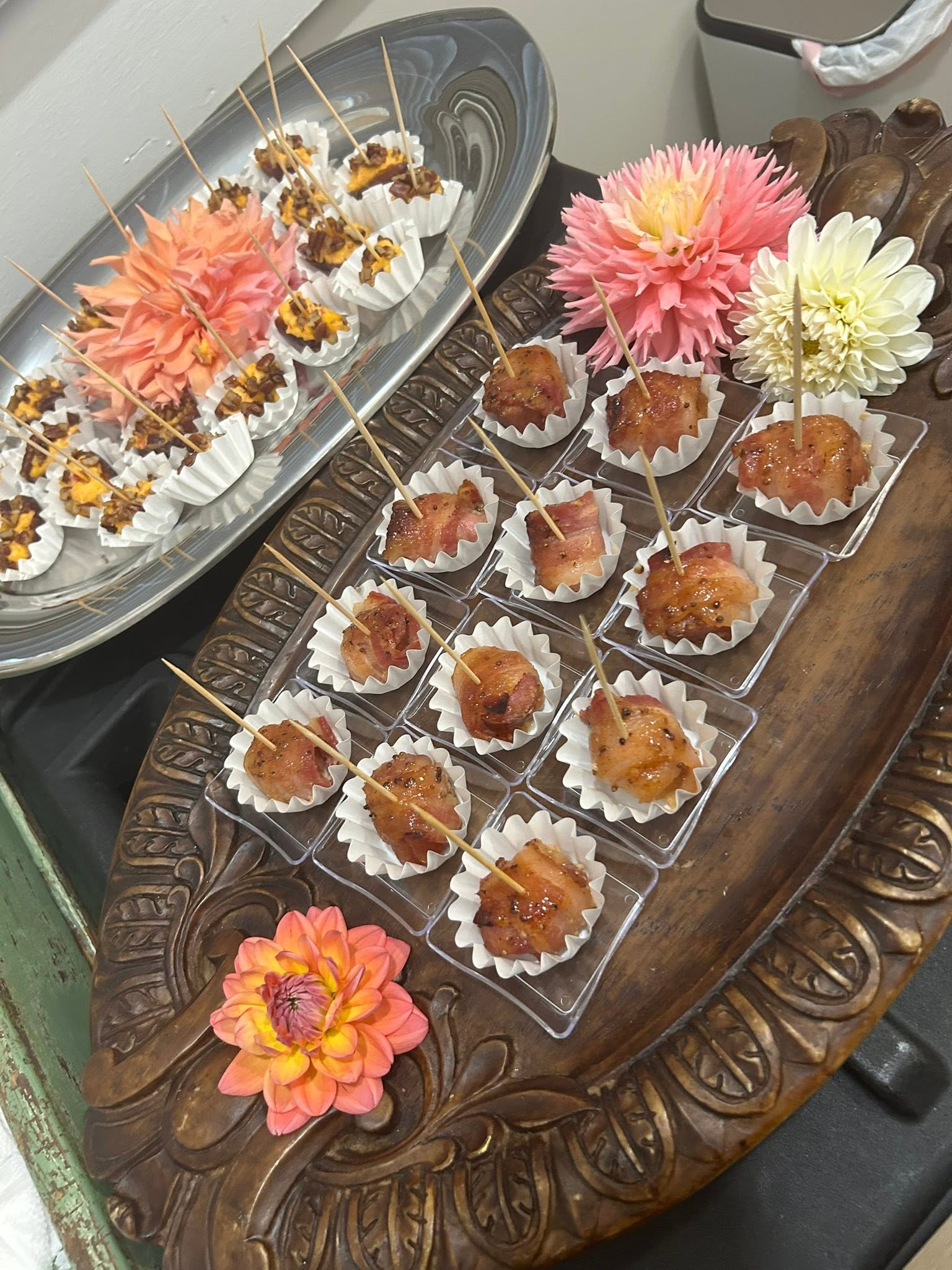Assorted bite-sized meat skewers in white paper cups arranged on a carved wooden tray with pink and yellow flowers nearby