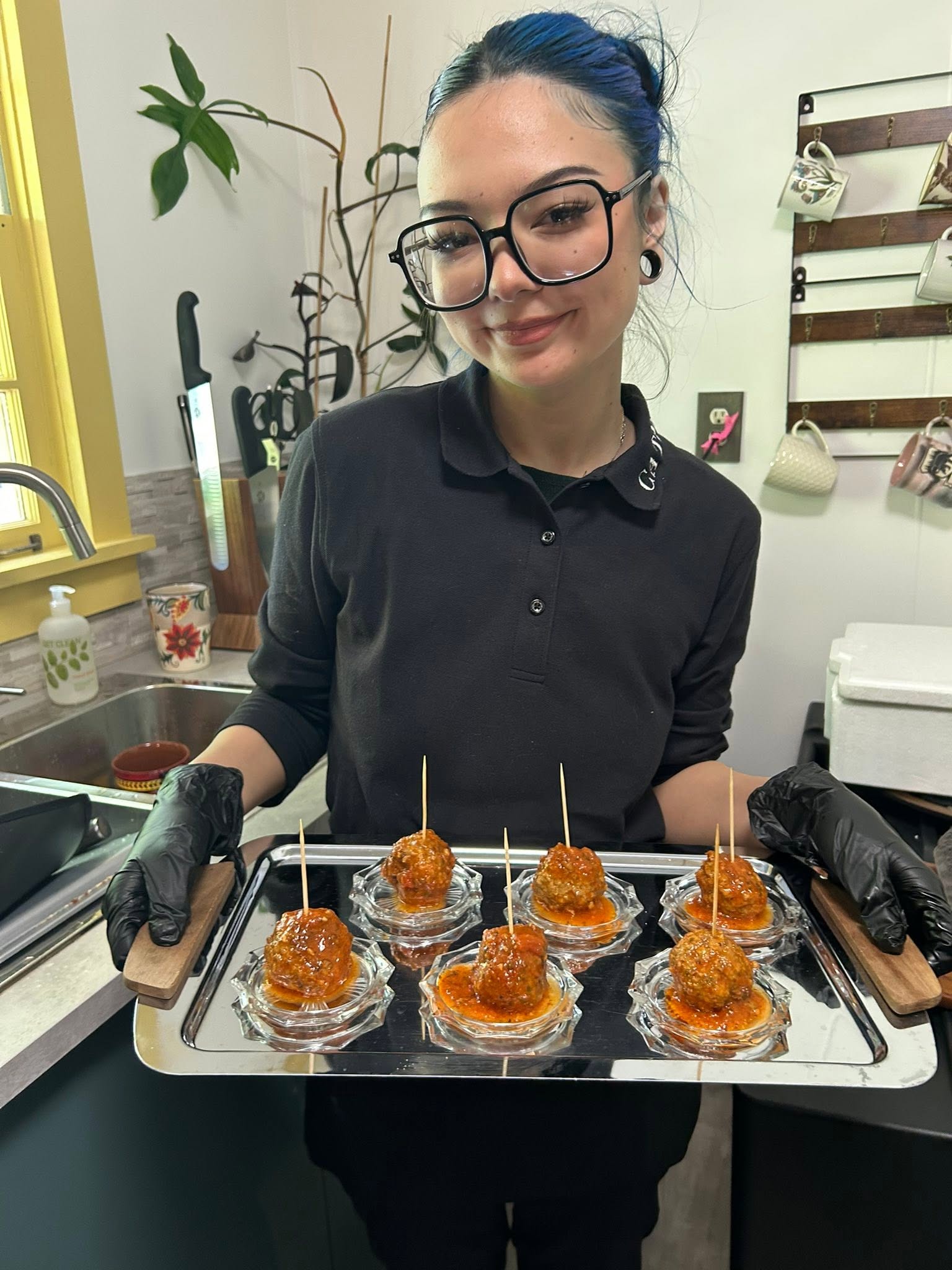A person with blue hair and large black glasses behind a kitchen counter presents a tray of glazed meatball skewers in aluminum cups.