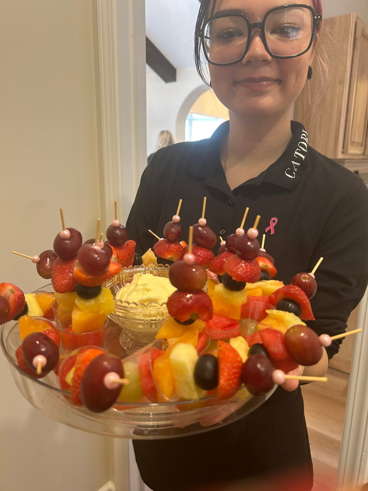 Catori Artisan Team Member Holding Fruit Skewers - CATORI Artisan Person wearing large black-rimmed glasses and a black collared shirt presents a large fruit-skewer platter with grapes, strawberries, and melon pieces.