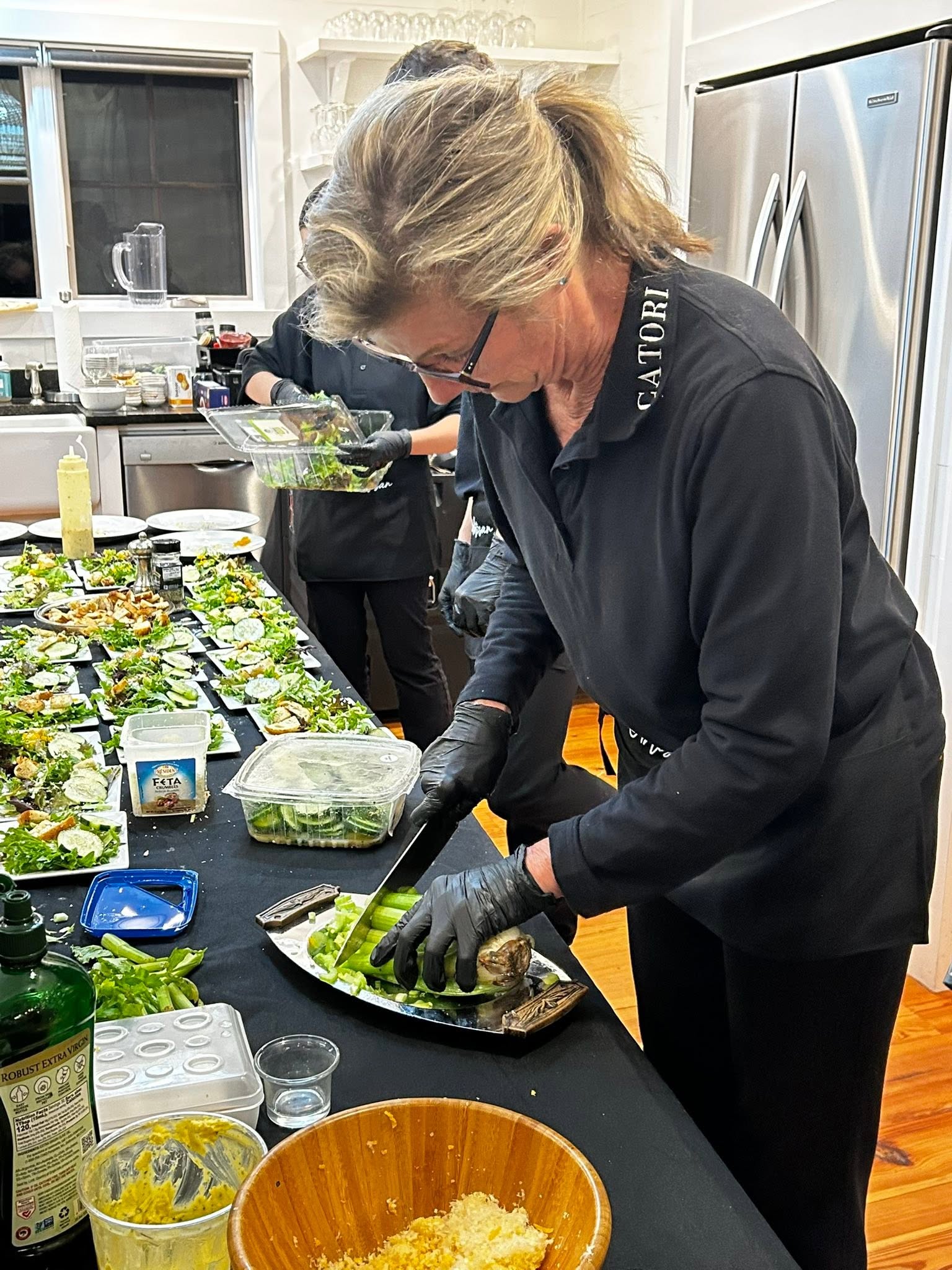 Cooks wearing black uniforms and gloves assemble small salads on a long serving table in a bright kitchen.