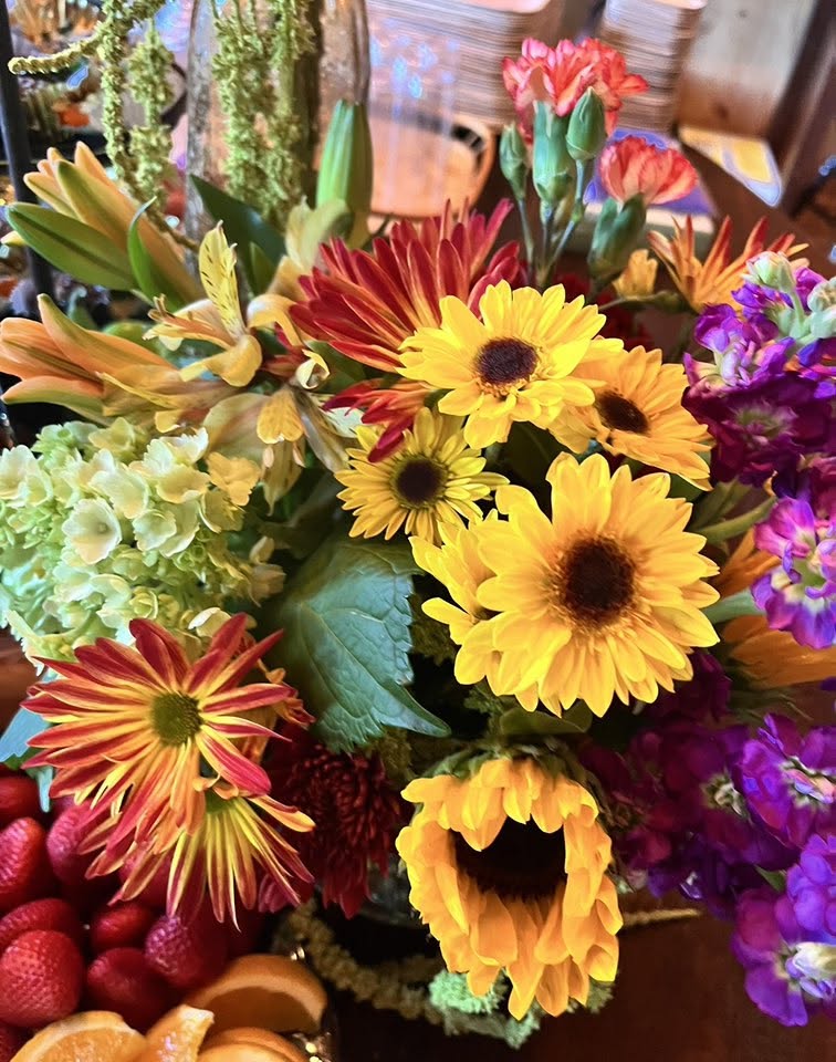 Catori Artisan Vibrant Event Flower Centerpiece - CATORI Artisan Vibrant bouquet featuring yellow sunflowers, orange-red mums, and purple flowers with green foliage on a table.