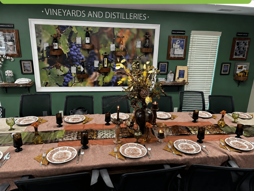 Catori Artisan Vineyard And Distillery Table Setting - CATORI Artisan Long dining table set for a formal meal in a green-walled room with a vineyard mural and decorative centerpiece of flowers and candles.