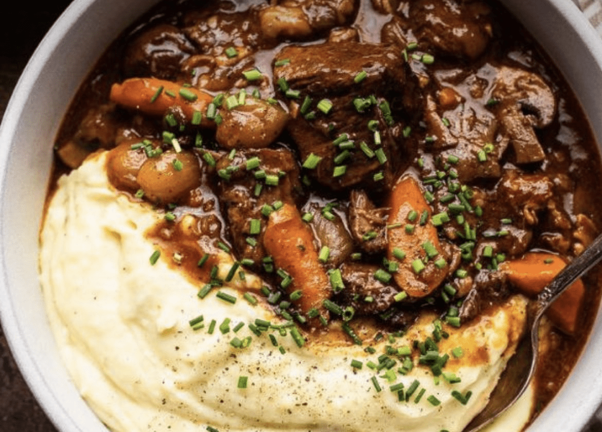 Beef stew with carrots and potatoes served over mashed potatoes, garnished with chopped chives in a shallow bowl