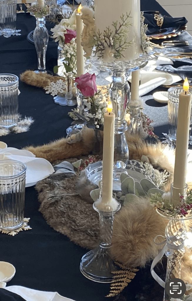 Elegant table centerpiece with tall ivory candles, glass vases, pink rose, dried grasses, and a furry brown runner on a dark table surface.