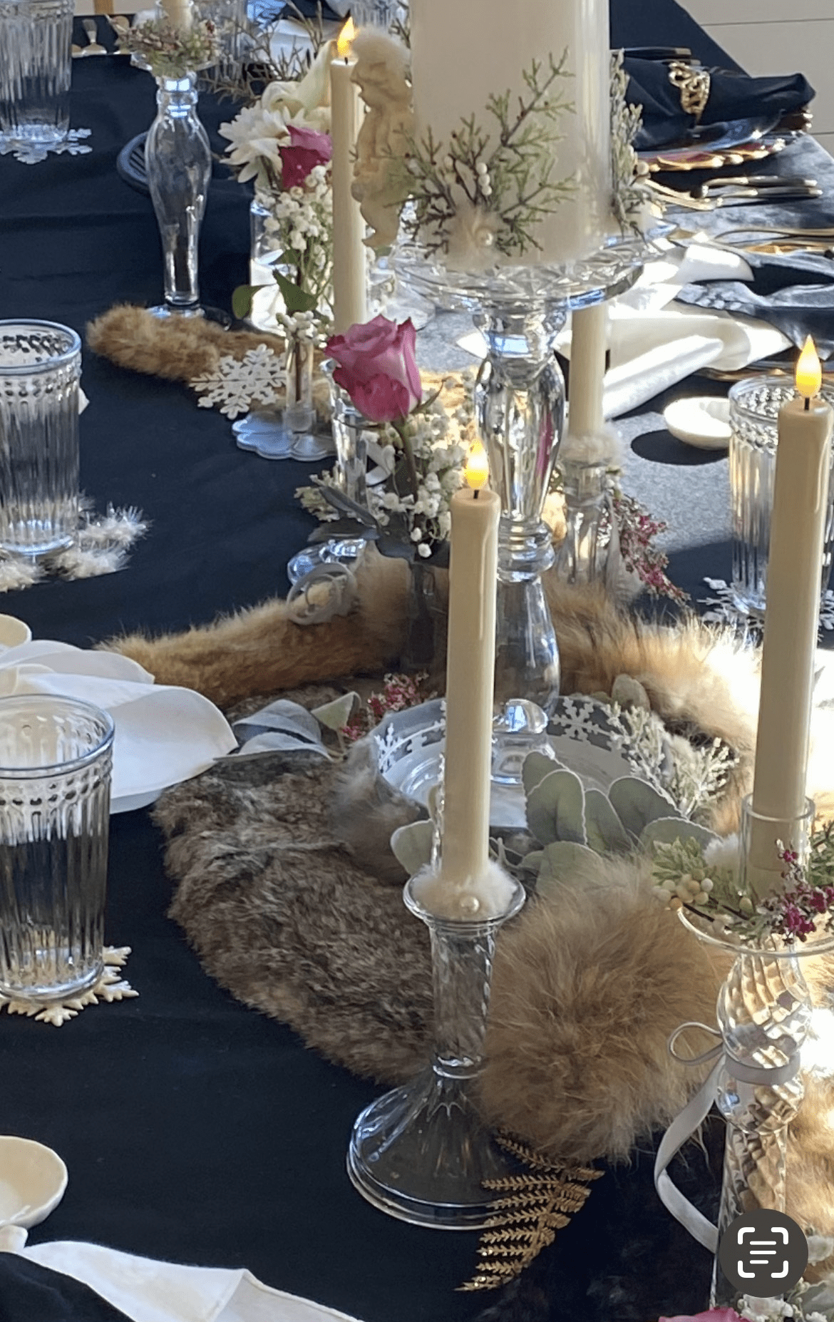Elegant table centerpiece with tall ivory candles in crystal holders, pink rose, white flowers, and fuzzy beige trim on a dark tablecloth.