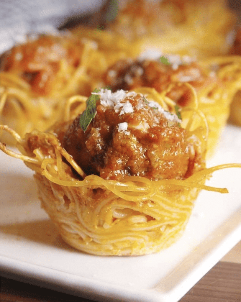 Spaghetti nest cup with a meatball, marinara sauce, grated cheese, and a basil leaf on top.