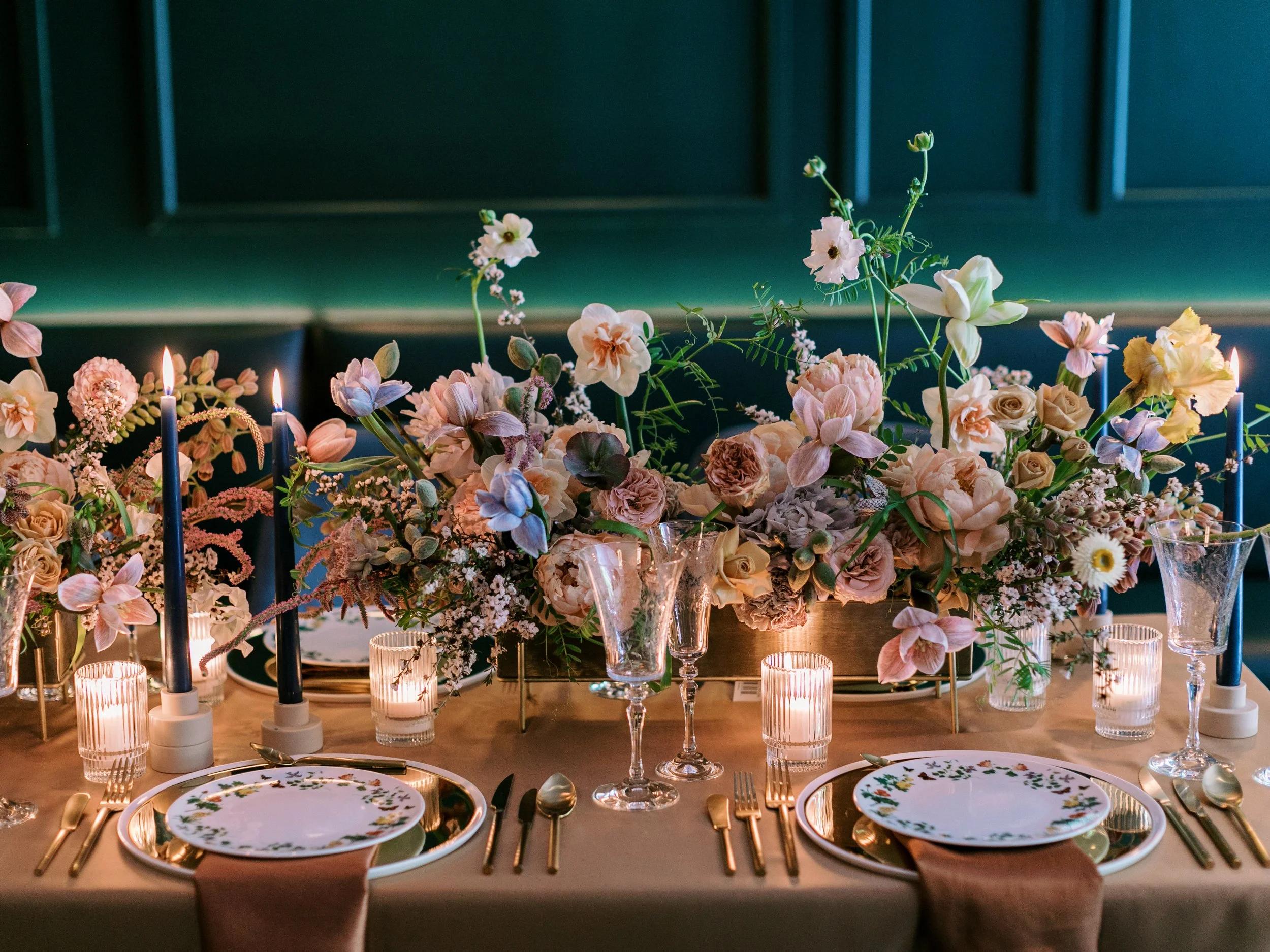 Elegant table setting with pastel flower centerpiece, blue taper candles, crystal wine glasses and gold-cutlery on a taupe tablecloth