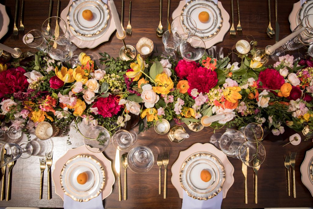 Top-down view of an elegant dining table set with gold flatware, ornate plates, candles, and a vibrant floral centerpiece.