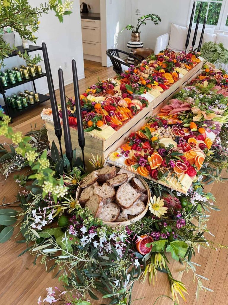 tast - CATORI Artisan Elaborate fruit and cheese platter arranged on a wooden tiered table, surrounded by a lush floral centerpiece and a bread basket.