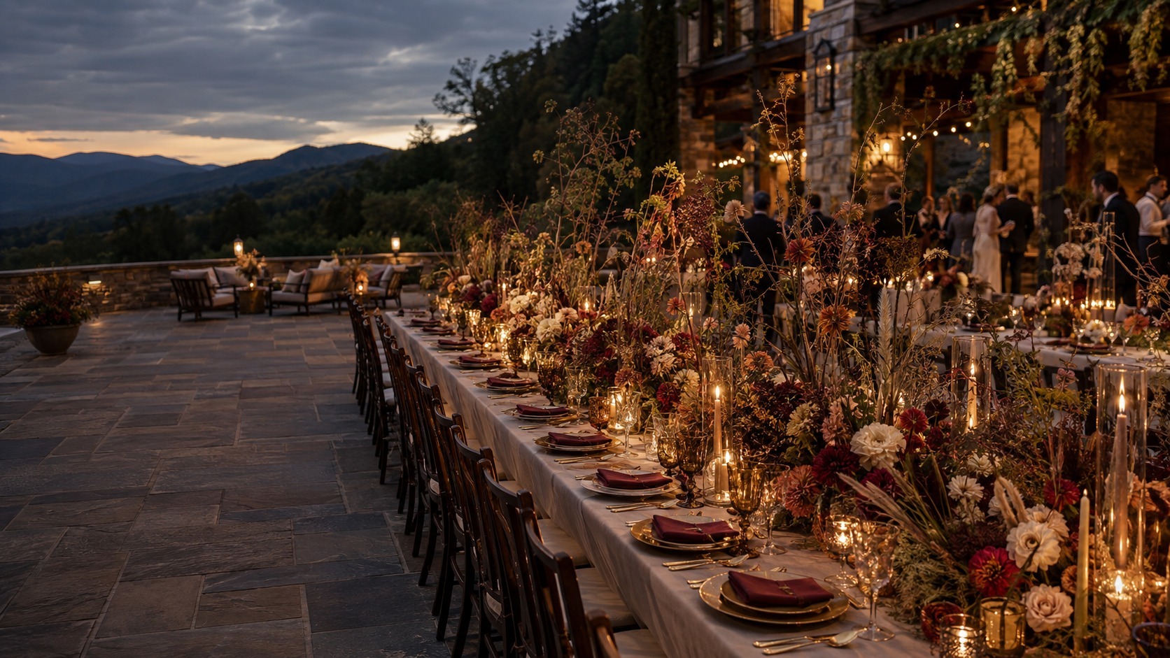 Long outdoor dining table at dusk with candlelight, burgundy napkins, gold-toned dinnerware, and lush floral arrangements; guests mingle in the background.