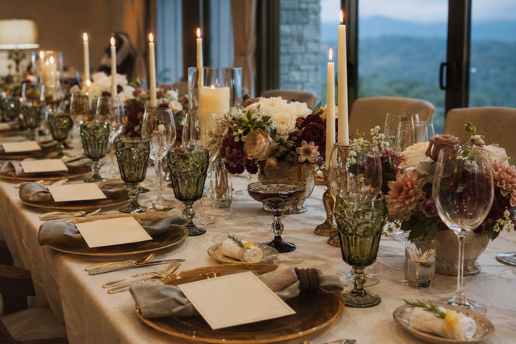 Elegant banquet table set for a formal dinner with gold charger plates, napkins, crystal and green-tinted goblets, and lush floral centerpieces under candlelight by a windowed backdrop.
